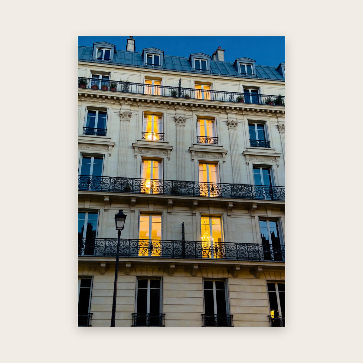 Affiche Paris à l'heure bleue : façade haussmannienne en pierre crème vue de face, balcons filants en fer forgé et fenêtres éclairées, un lustre