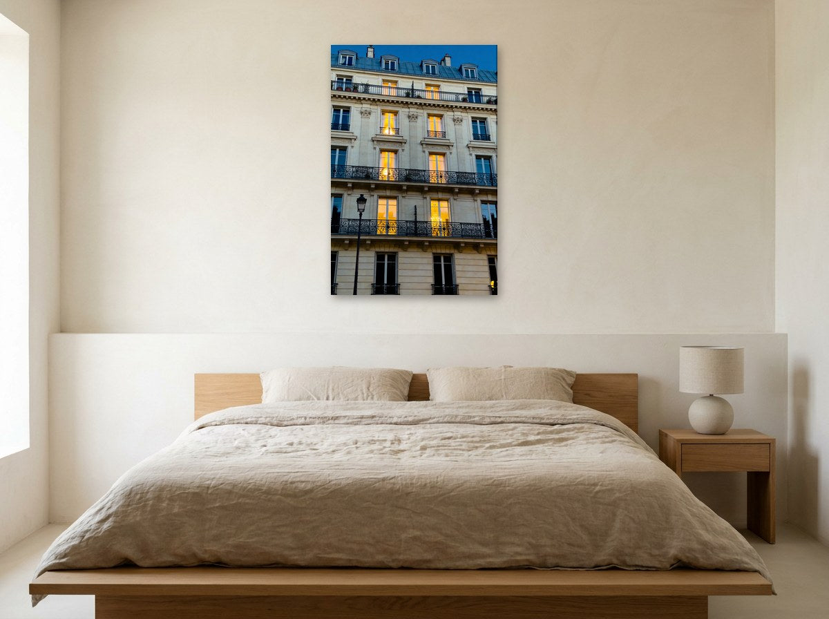 Mise en situation : toile des balcons filants au-dessus d'un lit en bois clair au linge de lin beige, sur le mur crème d'une chambre