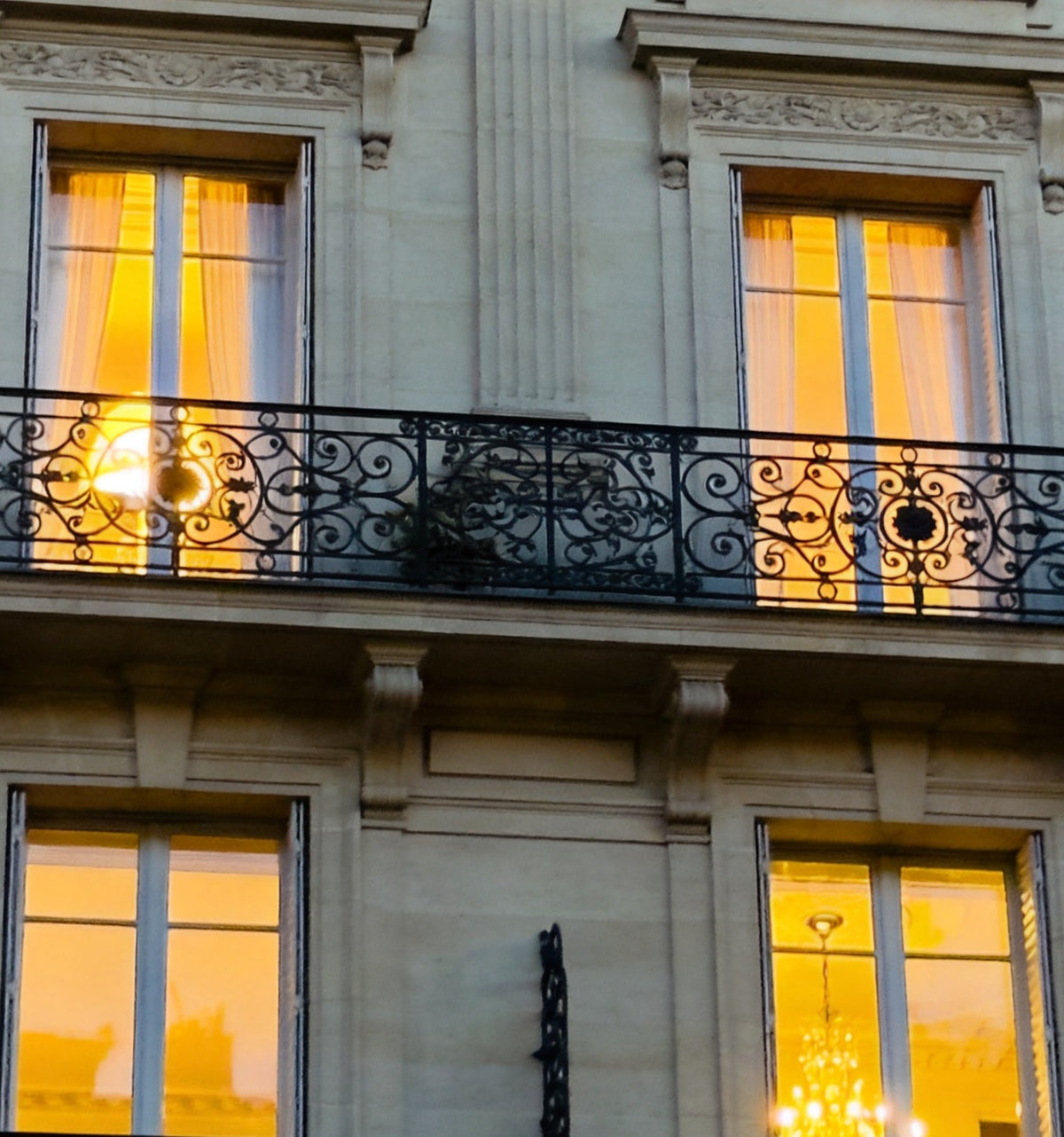 Détail : balcon en fer forgé à volutes devant des fenêtres éclairées aux voilages blancs