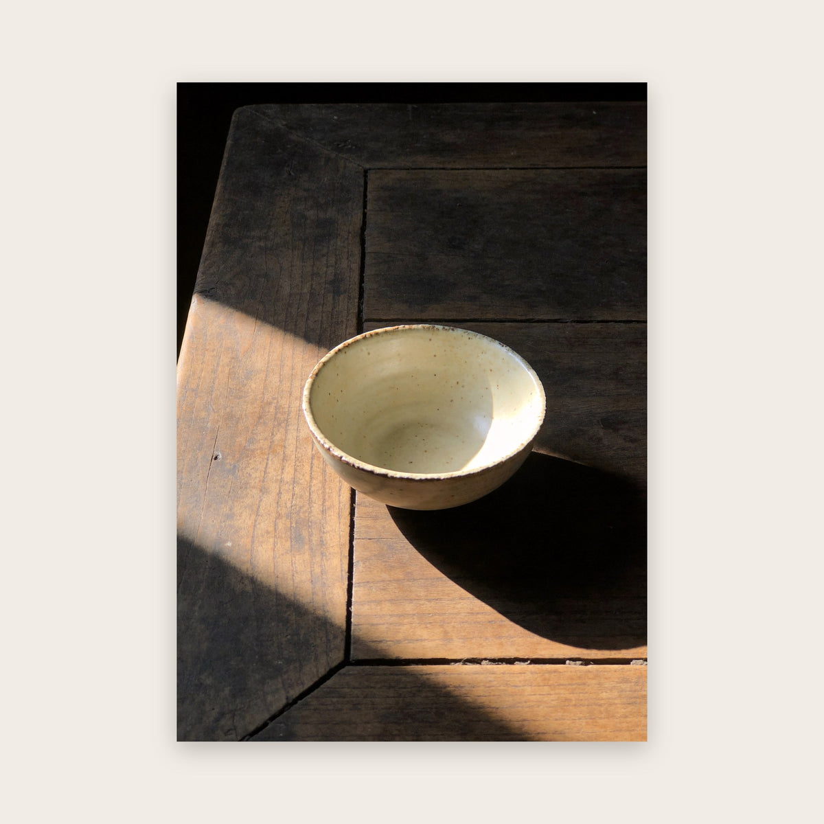 Affiche japandi au rendu photo : bol de céramique crème mouchetée sur une table de bois sombre, éclairé par un rai de soleil diagonal