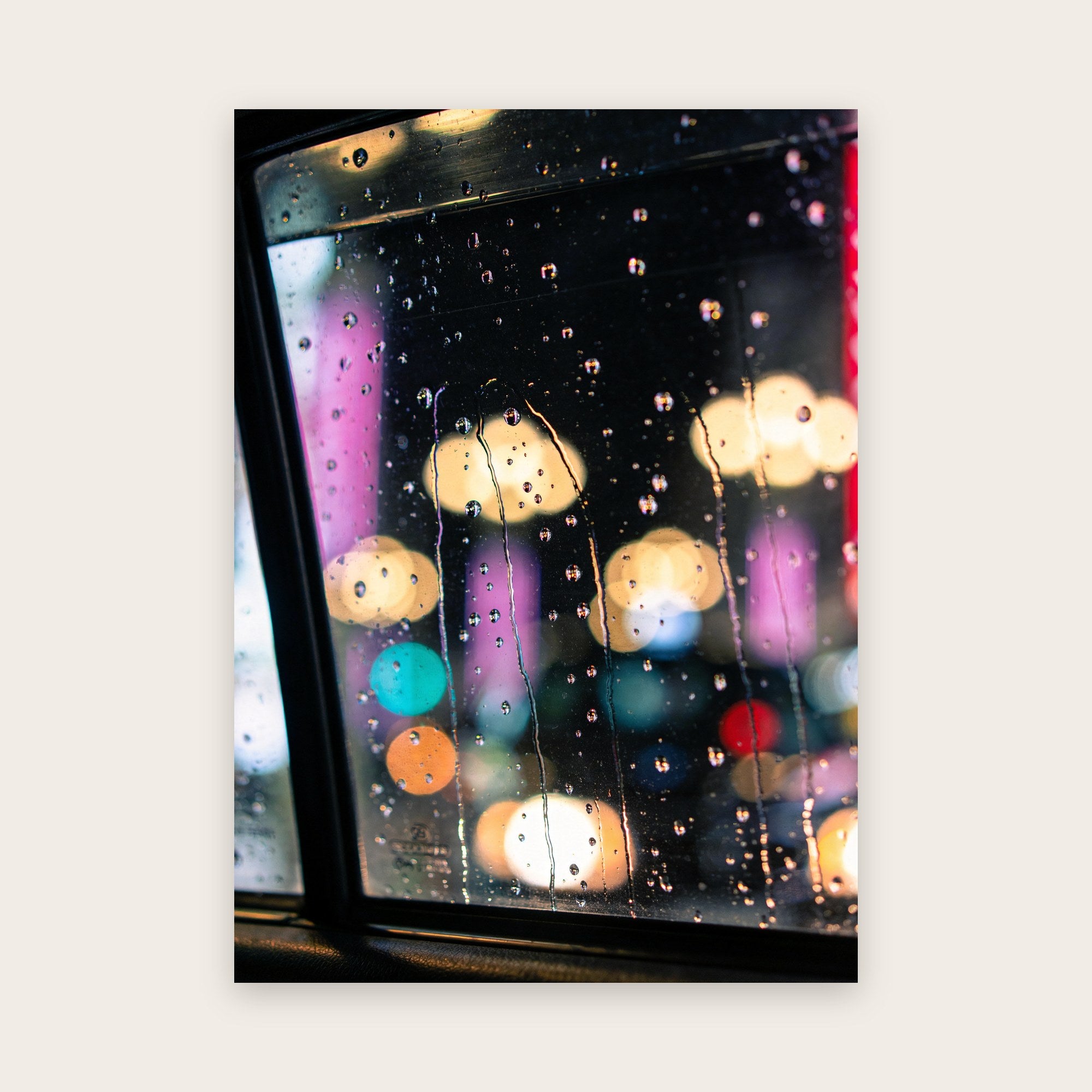 Vitre de voiture couverte de gouttes de pluie devant des halos flous crème, roses, violets et turquoise, la nuit à Tokyo